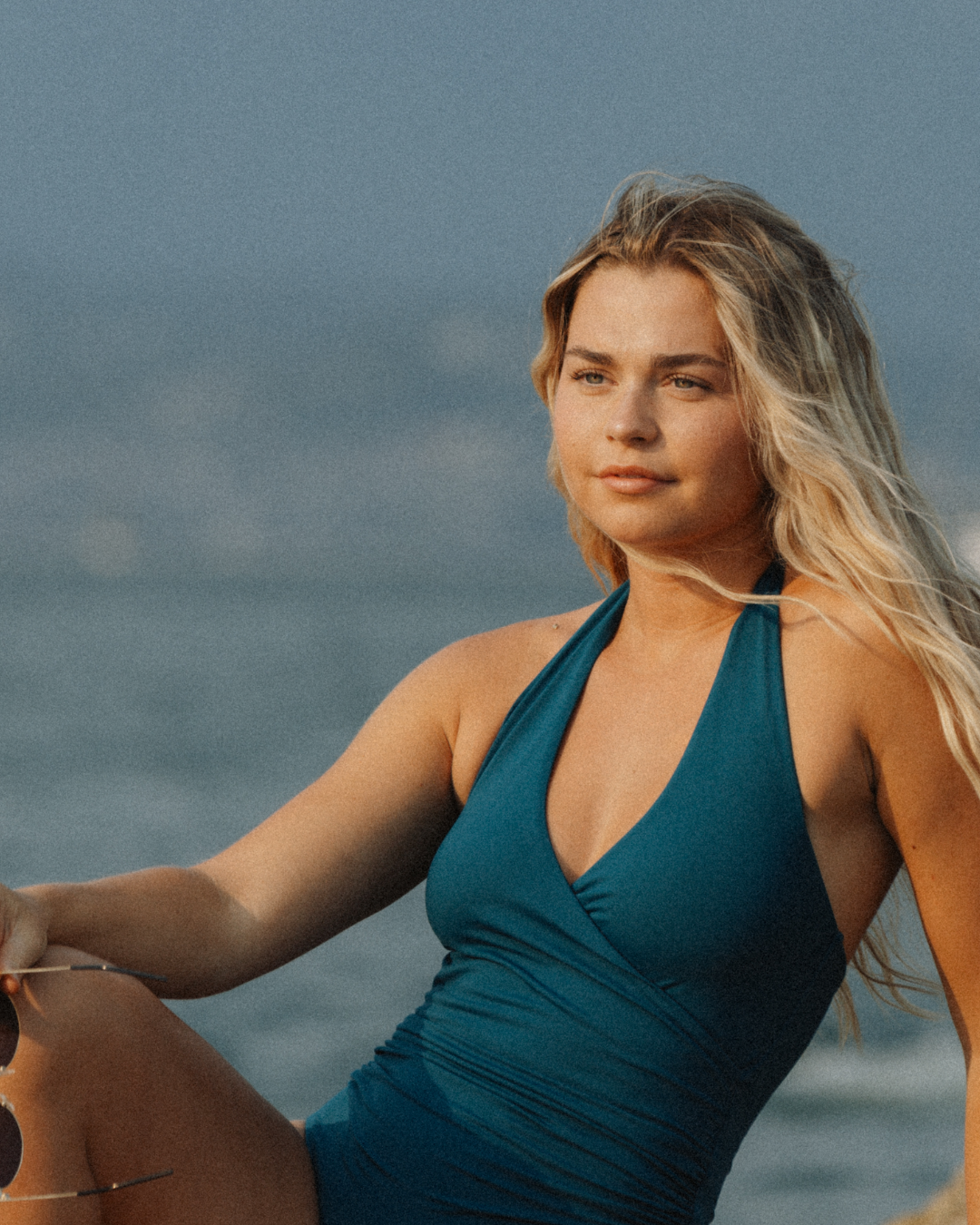 A woman sitting by the water wearing a green one-piece swimsuit with a halter neck and plunging v-neckline.