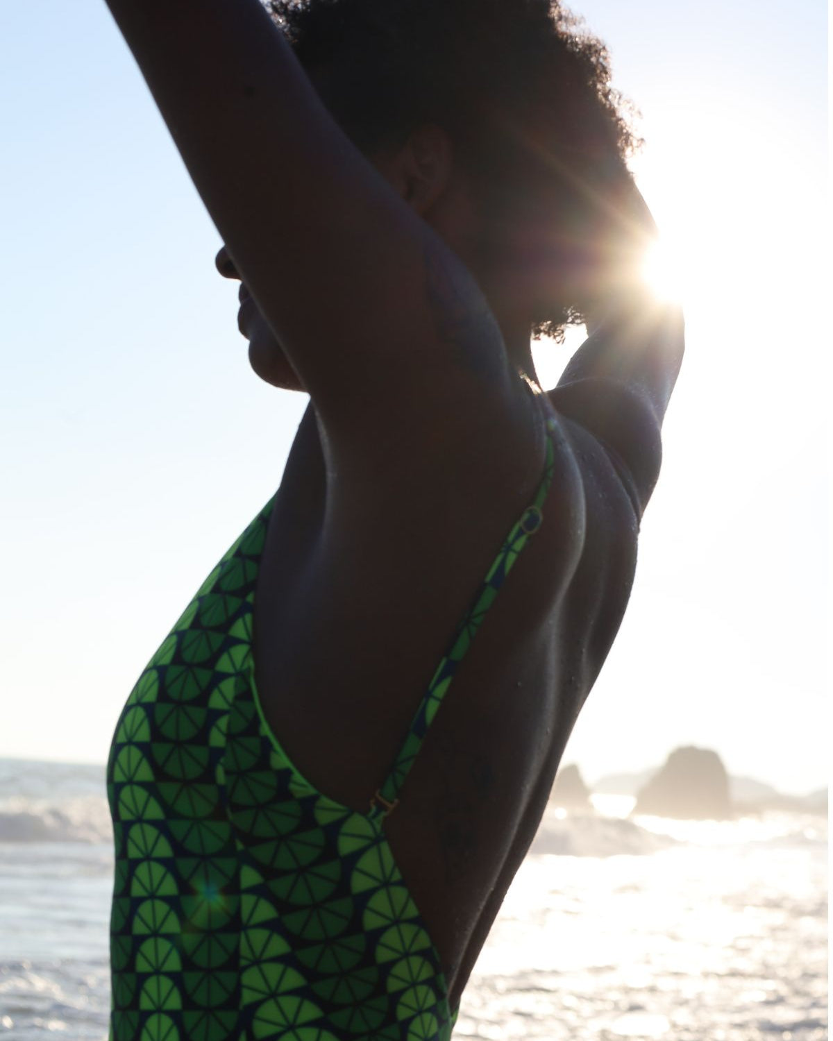 Person wearing a green and black patterned swimsuit with a blurred background of water and sky.