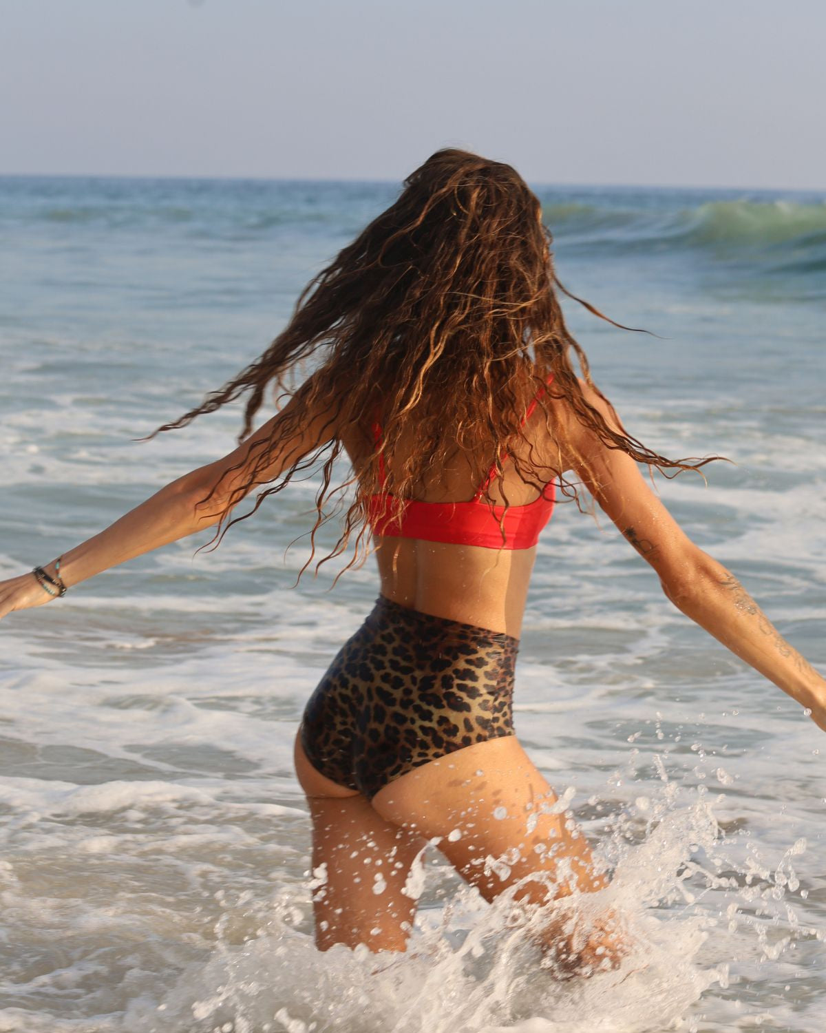 Person in a red bikini top and leopard print bottoms walking on a beach with ocean waves.