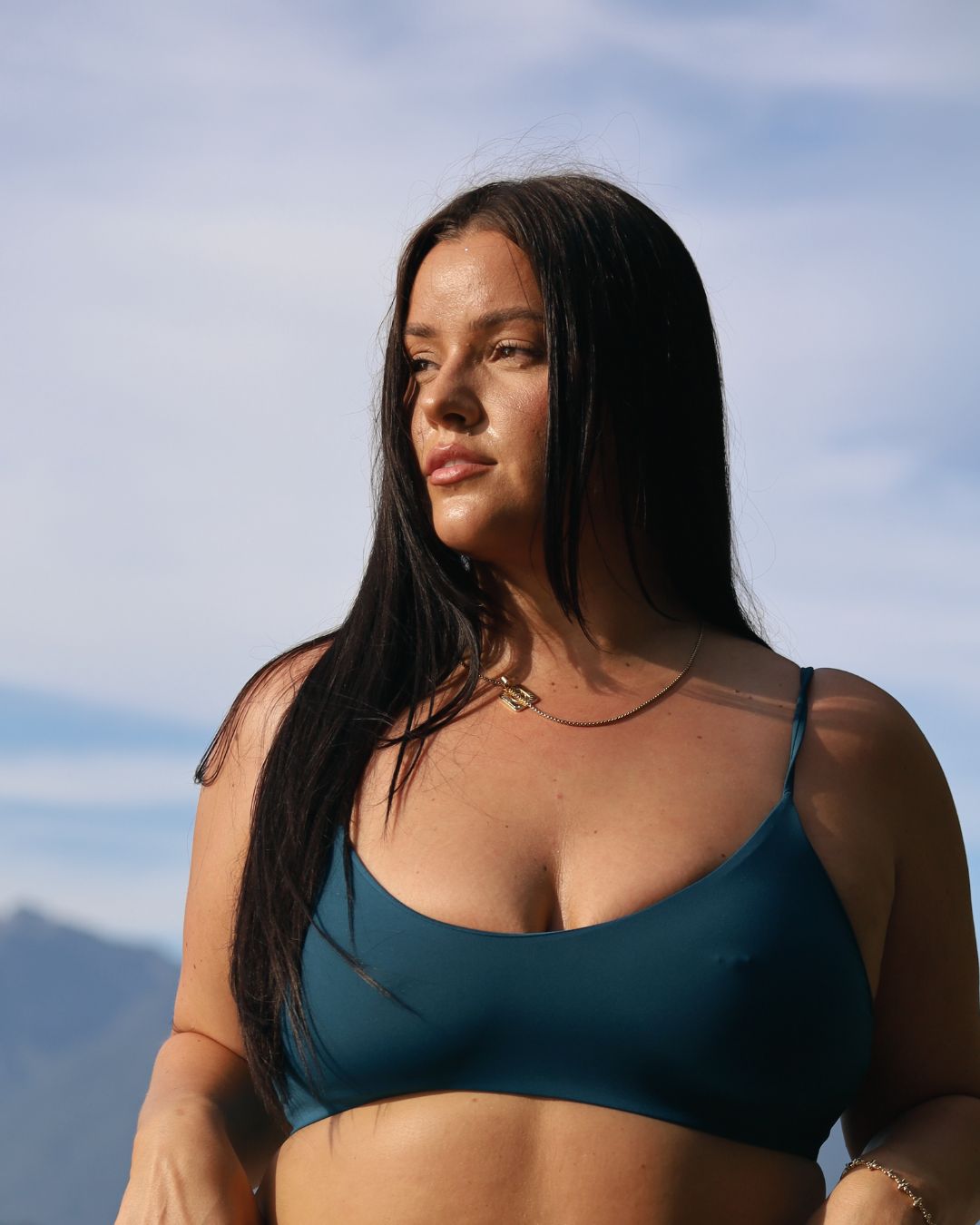 Woman wearing a teal bikini top against a blue sky and mountain background.A sustainable swimsuit from Canada.