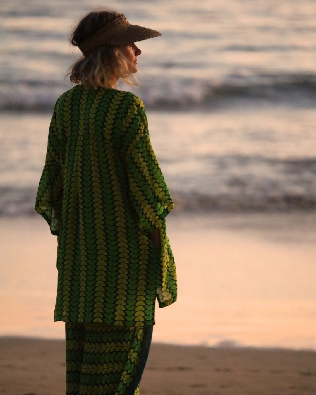 Person wearing a green and yellow patterned outfit standing on a beach at sunset.
