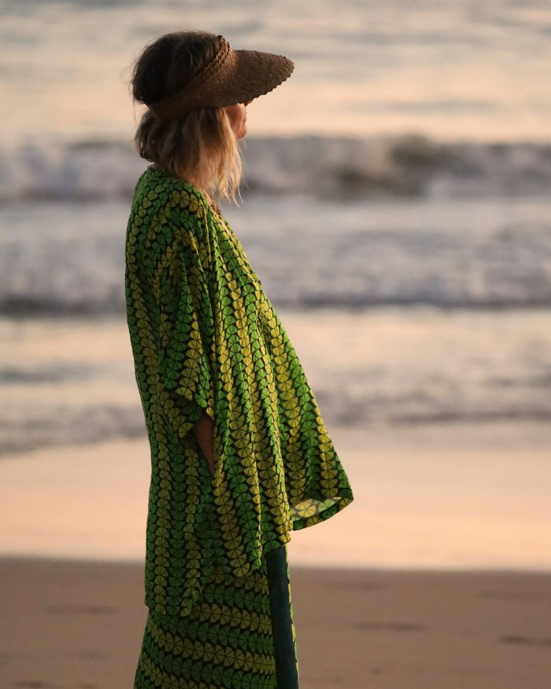 Woman wearing a green and yellow patterned dress and straw hat standing on a beach at sunset.